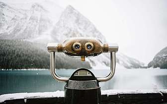 Fernrohr mit Regentropfen vor verschneitem Bergsee und bewaldeten Bergen