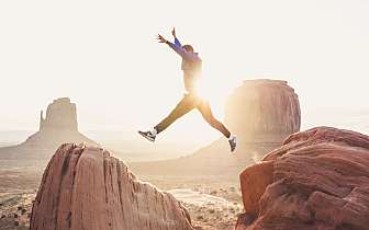 Person springt bei Sonnenuntergang zwischen zwei großen roten Felsen in einer Wüstenlandschaft